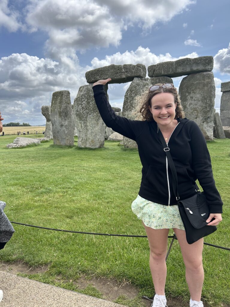 A girl wearing a black zip-up hoodie and green flowery skirt pretends she's holding up the stones at Stonehenge, with a huge dorky grin on her face.